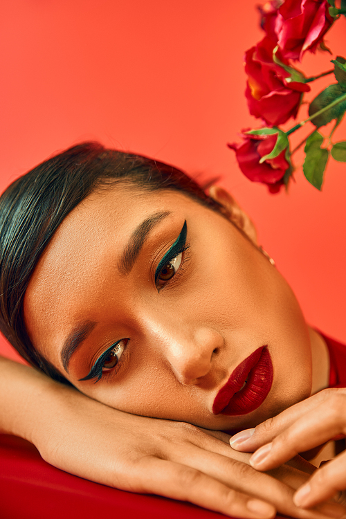 close up portrait of mesmerizing and pensive asian woman with bright makeup and brunette hair dreaming with hands near face next to flowers on red background, spring style, generation z