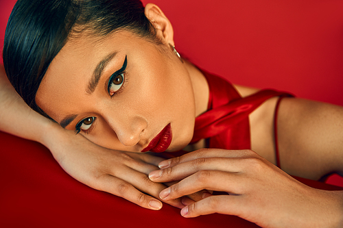 portrait of youthful and sensual asian woman with bold makeup and brunette hair laying on table and looking at camera on red background, generation z, spring fashion photography