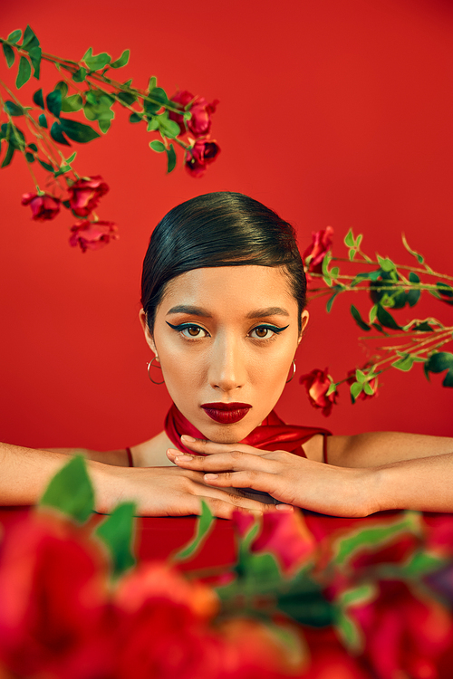 portrait of young and charming asian woman with bright makeup and brunette hair looking at camera surrounded with fresh roses on red background, spring fashion photography concept