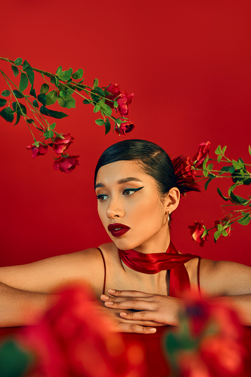 portrait of stylish and beautiful asian woman with bold makeup, brunette hair and neckerchief leaning on table and looking away near roses on red, blurred foreground