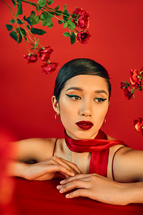 portrait of mesmerizing and dreamy asian woman posing near fresh roses on red background, bold makeup, brunette hair, stylish neckerchief, trendy spring concept