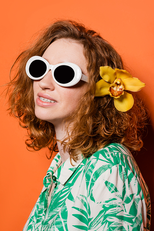 Young trendy red haired woman with sunglasses, blouse with floral print and orchid flower in hair smiling while standing on orange background, summer casual and fashion concept, Youth Culture