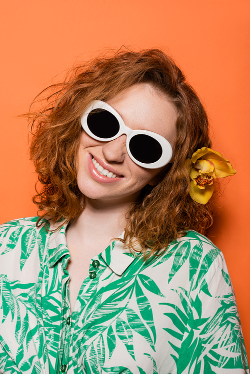Young redhead model with orchid flower in hair and sunglasses and blouse with floral pattern smiling and standing on orange background, summer casual and fashion concept, Youth Culture