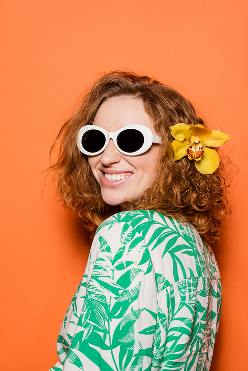 Positive young woman with red hair and orchid flower posing in sunglasses and blouse with floral print on orange background, summer casual and fashion concept, Youth Culture
