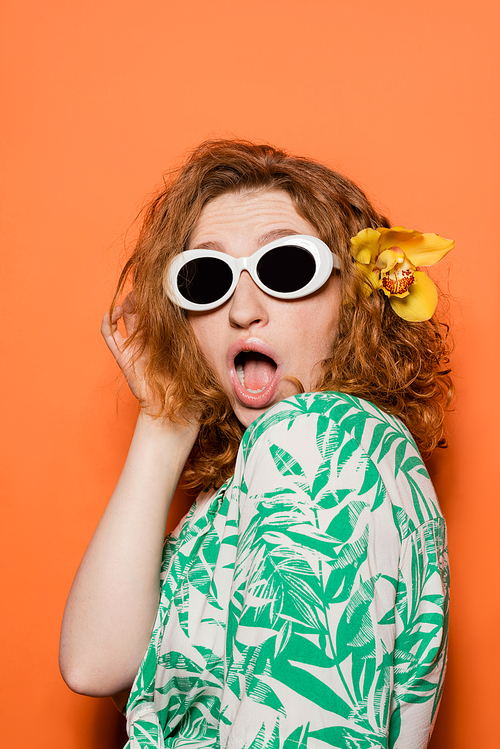 Shocked young redhead woman with orchid flower and sunglasses posing in blouse with floral pattern and standing on orange background, summer casual and fashion concept, Youth Culture