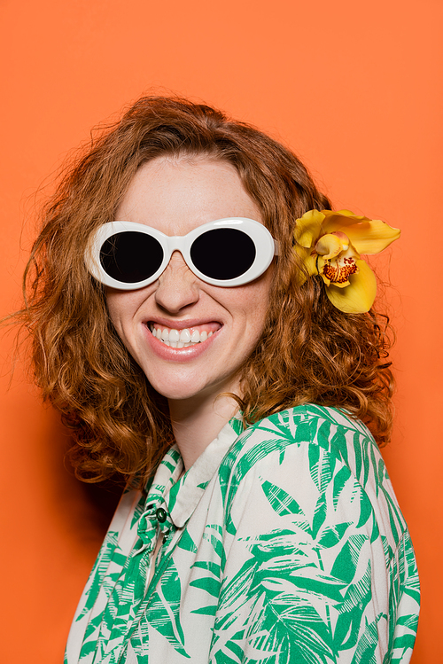 Positive woman with orchid flower in red hair wearing sunglasses and blouse with floral pattern while posing and standing on orange background, summer casual and fashion concept, Youth Culture