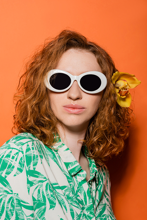 Portrait of stylish redhead woman with orchid flower in hair posing in sunglasses and blouse with floral pattern on orange background, summer casual and fashion concept, Youth Culture