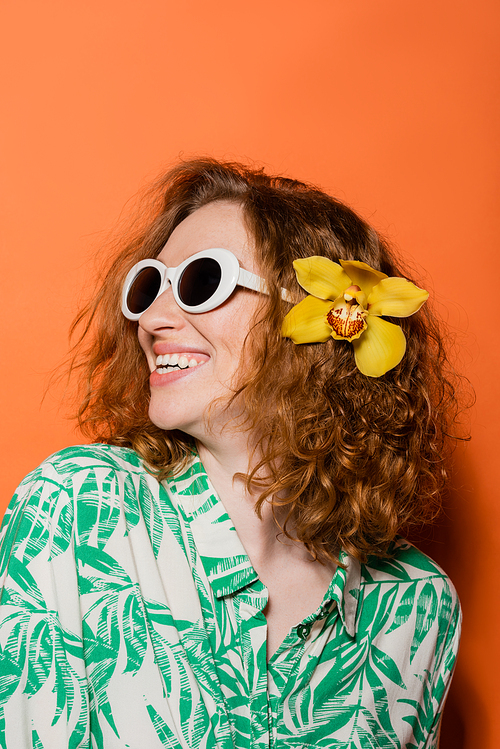 Happy and confident young redhead woman with orchid flower in hair, stylish sunglasses and modern blouse standing on orange background, summer casual and fashion concept, Youth Culture