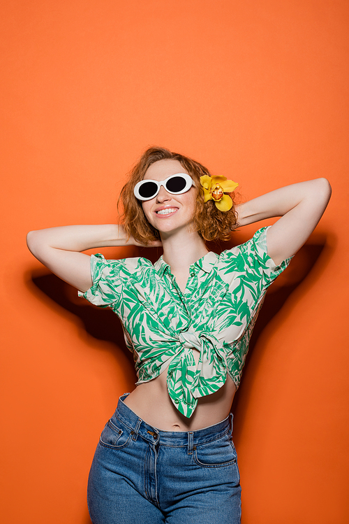 Joyful young redhead woman with orchid flower in hair and sunglasses posing in blouse with floral pattern and jeans on orange background, summer casual and fashion concept, Youth Culture