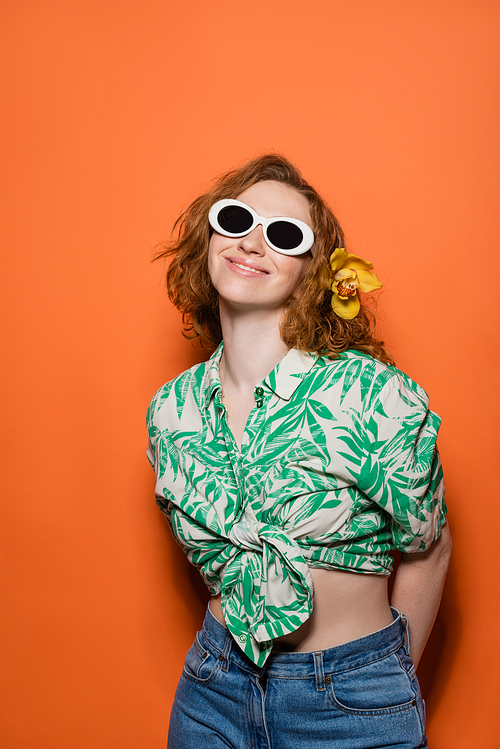 Positive young woman with orchid flower in red hair and sunglasses wearing jeans and blouse with floral print on orange background, summer casual and fashion concept, Youth Culture