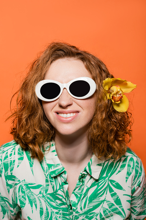 Smiling young woman with orchid flower in red hair wearing sunglasses and blouse with floral print while standing isolated on orange, stylish casual outfit and summer vibes concept, Youth Culture