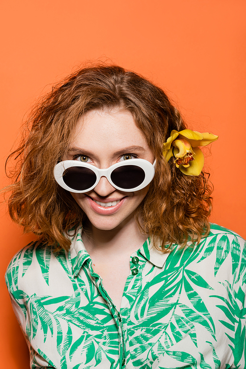 Portrait of positive young woman with orchid flower in red hair and sunglasses looking at camera while standing on orange background, summer casual and fashion concept, Youth Culture