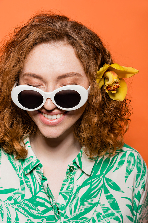Joyful red haired woman with sunglasses and orchid flower wearing blouse with floral print and standing isolated on orange, stylish casual outfit and summer vibes concept, Youth Culture