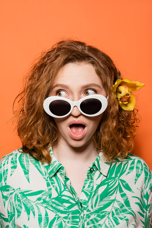 Shocked young woman with orchid flower in red hair wearing sunglasses and looking away while posing and standing on orange background, summer casual and fashion concept, Youth Culture