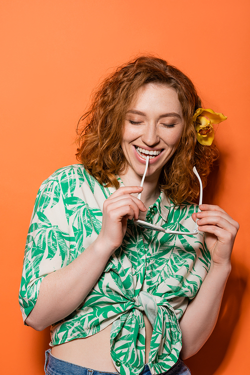 Positive redhead woman with orchid flower holding fashionable sunglasses and closing eyes while standing on orange background, summer casual and fashion concept, Youth Culture