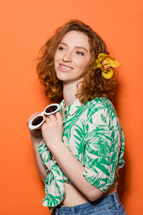 Smiling young redhead woman with orchid flower in hair wearing blouse with floral print and holding stylish sunglasses on orange background, summer casual and fashion concept, Youth Culture