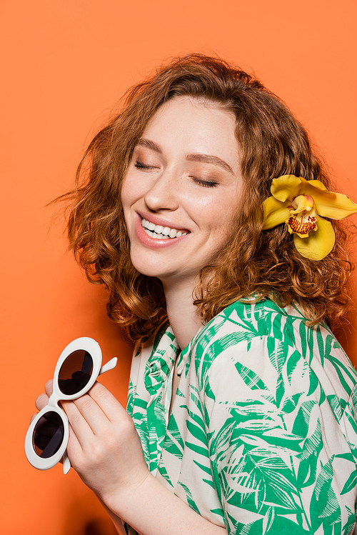 Positive young redhead model with orchid flower in hair holding sunglasses and wearing blouse with modern floral pattern on orange background, summer casual and fashion concept, Youth Culture