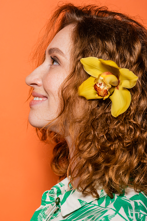 Side view of smiling young woman with orchid flower in red hair and natural makeup looking away while standing on orange background, summer casual and fashion concept, Youth Culture