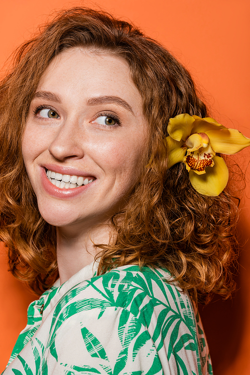Portrait of joyful young and red haired woman with orchid flower in hair looking away and posing in blouse while standing on orange background, summer casual and fashion concept, Youth Culture