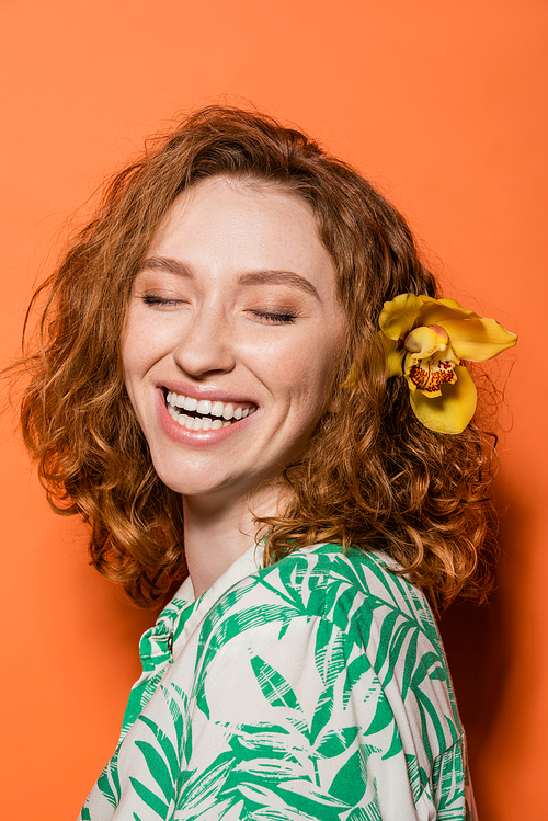 Happy young woman with orchid flower in red hair and trendy blouse with floral print standing with closed eyes on orange background, summer casual and fashion concept, Youth Culture