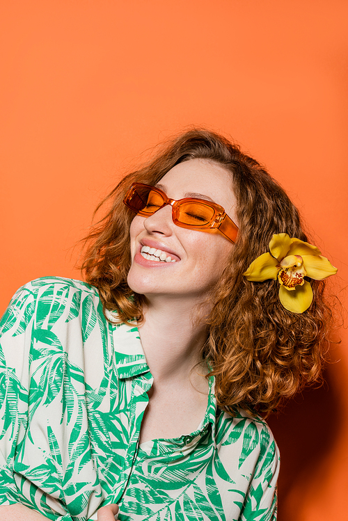 Positive young woman with orchid flower in red hair wearing sunglasses and stylish blouse while standing on orange background, summer casual and fashion concept, Youth Culture