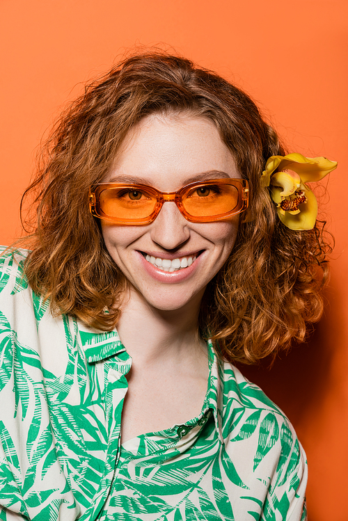 Portrait of smiling and stylish young woman with orchid flower in red hair looking at camera and posing in sunglasses on orange background, summer casual and fashion concept, Youth Culture