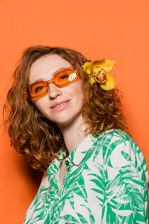 Positive young model with orchid flower in red hair, natural makeup and sunglasses posing in blouse with floral print on orange background, summer casual and fashion concept, Youth Culture