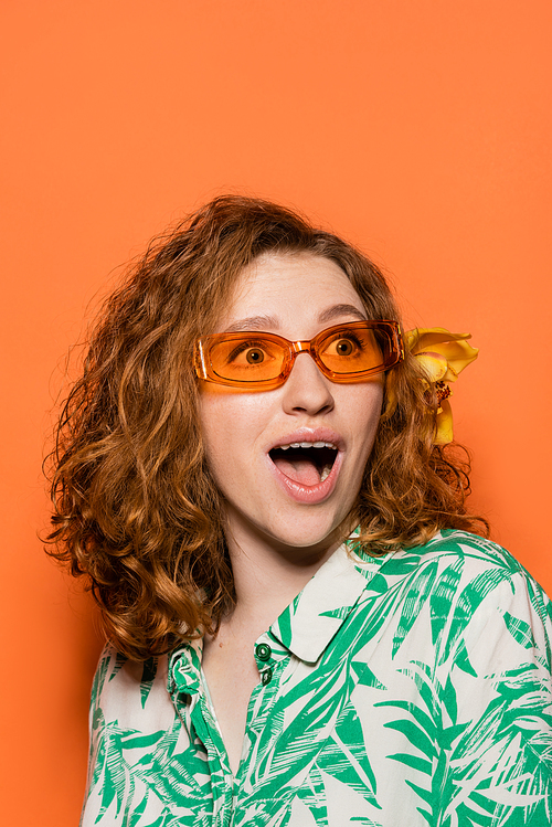 Excited young redhead woman with orchid flower in hair and sunglasses looking away while wearing blouse with floral pattern on orange background, summer casual and fashion concept, Youth Culture