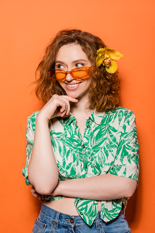 Cheerful young woman with orchid flower in hair wearing sunglasses and blouse with floral pattern while standing on orange background, summer casual and fashion concept, Youth Culture