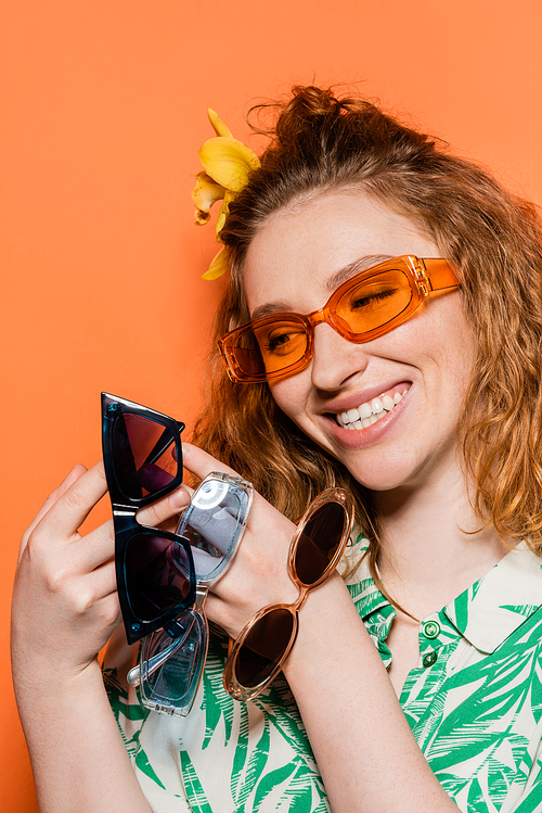 Cheerful young redhead woman with orchid flower in hair wearing and holding modern sunglasses while standing isolated on orange, stylish casual outfit and summer vibes concept, Youth Culture