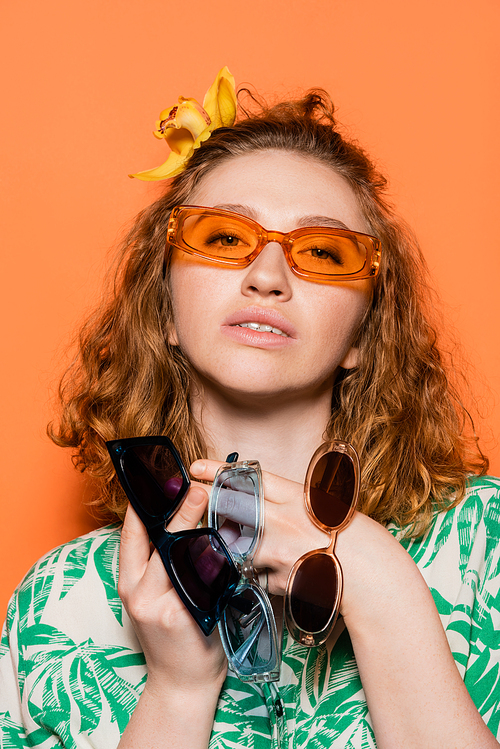 Portrait of stylish young woman with orchid flower in red hair holding sunglasses and looking at camera while standing on orange background, summer casual and fashion concept, Youth Culture