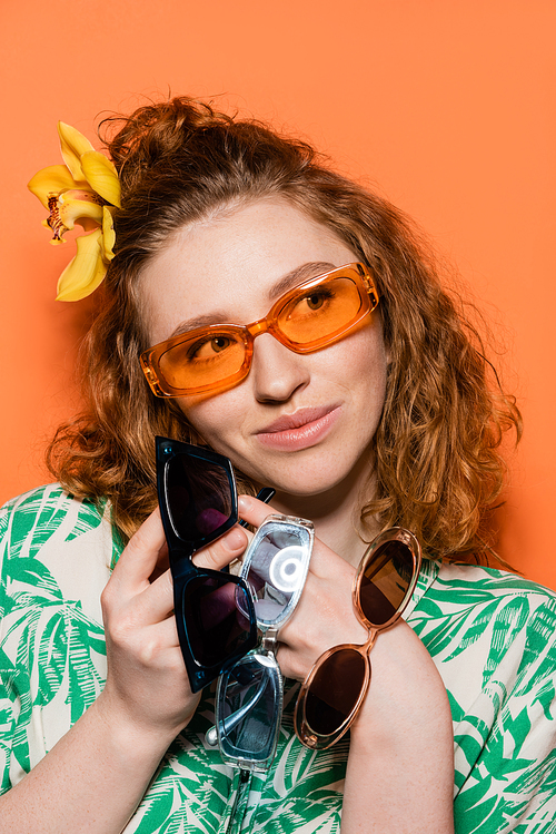 Portrait of joyful young woman with orchid flower in hair holding sunglasses and looking away while posing and standing on orange background, summer casual and fashion concept, Youth Culture