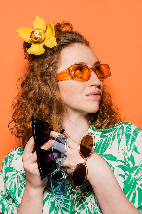 Trendy young woman with orchid flower in hair holding sunglasses and looking away while posing and standing on orange background, summer casual and fashion concept, Youth Culture
