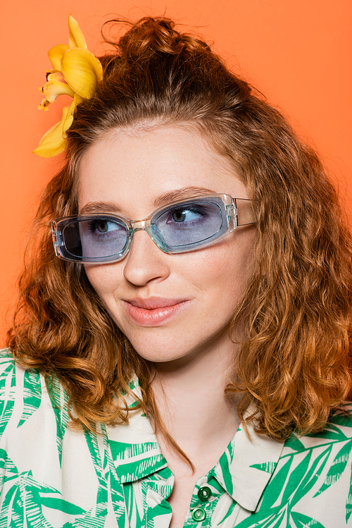 Portrait of young redhead model with orchid flower in hair wearing blue sunglasses and trendy blouse with floral pattern isolated on orange, stylish casual outfit and summer vibes concept