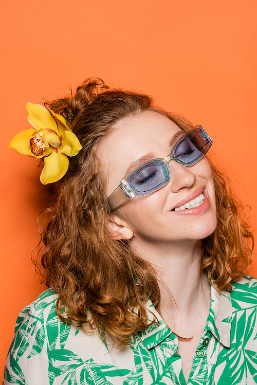 Smiling and trendy young woman with red hair and orchid flower posing in sunglasses and blouse with floral print on orange background, summer casual and fashion concept, Youth Culture