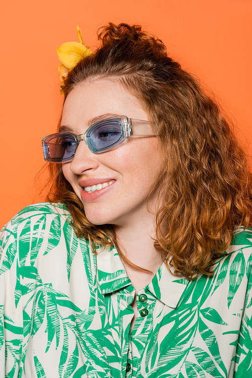 Portrait of smiling redhead woman with flower in hair and sunglasses posing in blouse with floral pattern isolated on orange, stylish casual outfit and summer vibes concept, Youth Culture