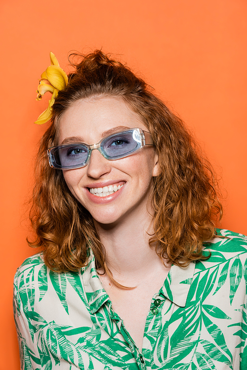 Cheerful young redhead woman with orchid flower in hair and blue sunglasses looking at camera while standing isolated on orange, stylish casual outfit and summer vibes concept