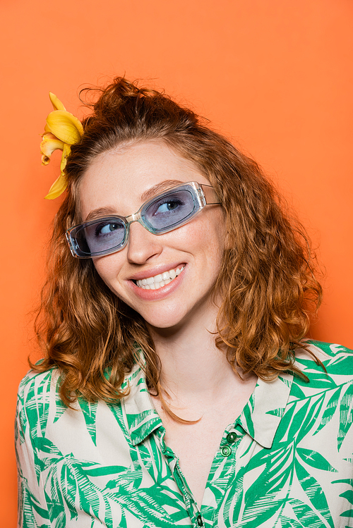 Portrait of joyful young redhead woman with orchid flower in hair and blue sunglasses looking away and standing on orange background, summer casual and fashion concept