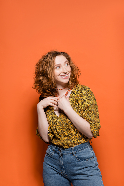 Cheerful and stylish young redhead woman in casual modern blouse and jeans touching necklaces and standing on orange background, stylish casual outfit and summer vibes concept, Youth Culture