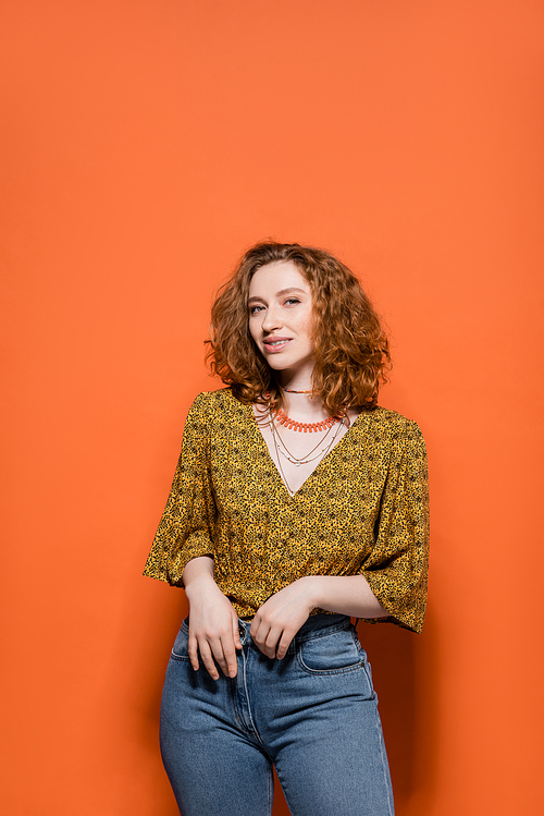 Trendy young redhead model in yellow blouse with abstract pattern and jeans smiling at camera and standing on orange background, stylish casual outfit and summer vibes concept, Youth Culture