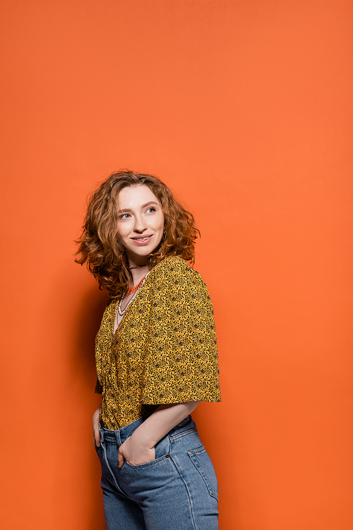 Joyful young redhead woman in blouse with abstract pattern holding hands in pockets of jeans and looking away on orange background, stylish casual outfit and summer vibes concept, Youth Culture