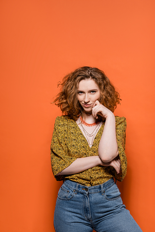Confident and smiling red haired woman in blouse with abstract pattern and jeans looking at camera while standing on orange background, stylish casual outfit and summer vibes concept, Youth Culture
