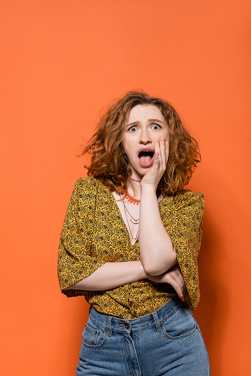 Shocked young redhead woman in blouse with abstract pattern and jeans looking at camera and standing on orange background, stylish casual outfit and summer vibes concept, Youth Culture