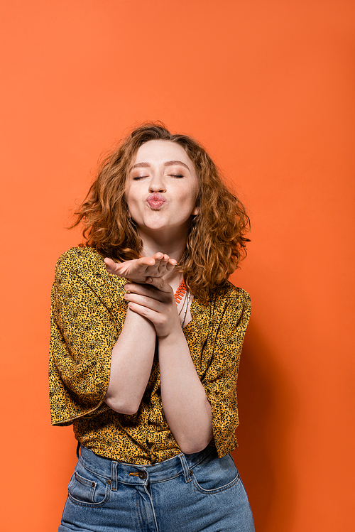 Young and stylish red haired woman in yellow blouse with abstract pattern and jeans blowing air kiss at camera on orange background, stylish casual outfit and summer vibes concept, Youth Culture