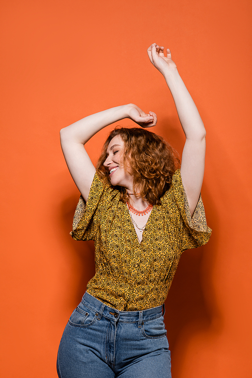 Cheerful young red haired woman in yellow blouse with abstract pattern and jeans dancing while standing on orange background, stylish casual outfit and summer vibes concept, Youth Culture