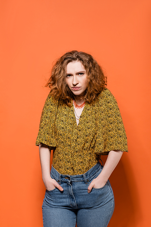 Serious young red haired model in blouse with abstract print and jeans holing hands in pockets and looking at camera on orange background, stylish casual outfit and summer vibes concept