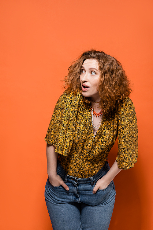 Shocked young red haired woman in yellow blouse and trendy jeans looking away while posing and standing on orange background, stylish casual outfit and summer vibes concept, Youth Culture