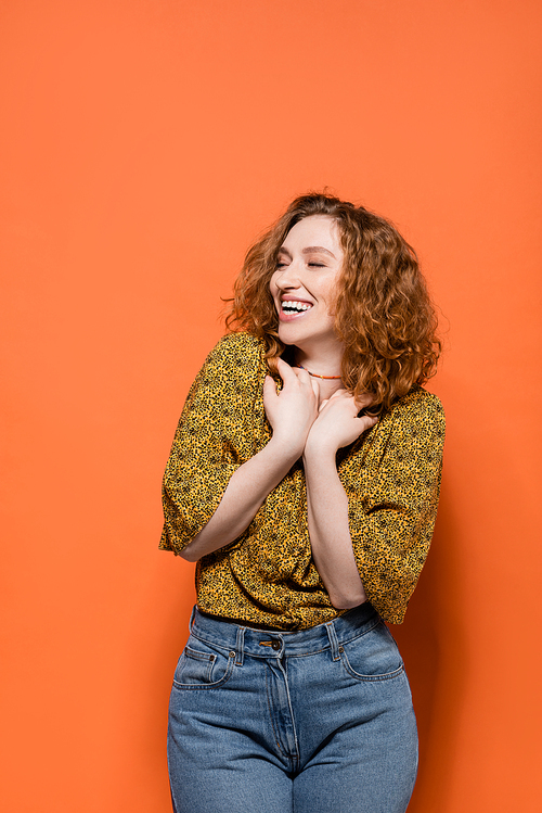 Joyful and stylish young redhead woman in yellow blouse and jeans looking away while standing on orange background, stylish casual outfit and summer vibes concept, Youth Culture