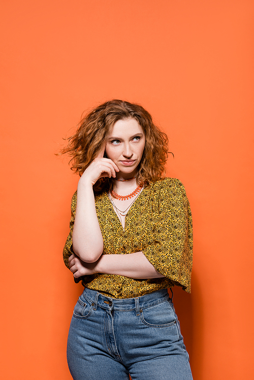 Pensive young red haired woman in modern yellow blouse and jeans looking away while standing and posing on orange background, stylish casual outfit and summer vibes concept, Youth Culture