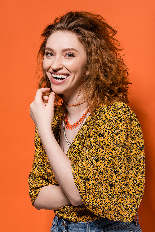 Portrait of young red haired woman in blouse with abstract print and jeans laughing and looking at camera on orange background, stylish casual outfit and summer vibes concept, Youth Culture